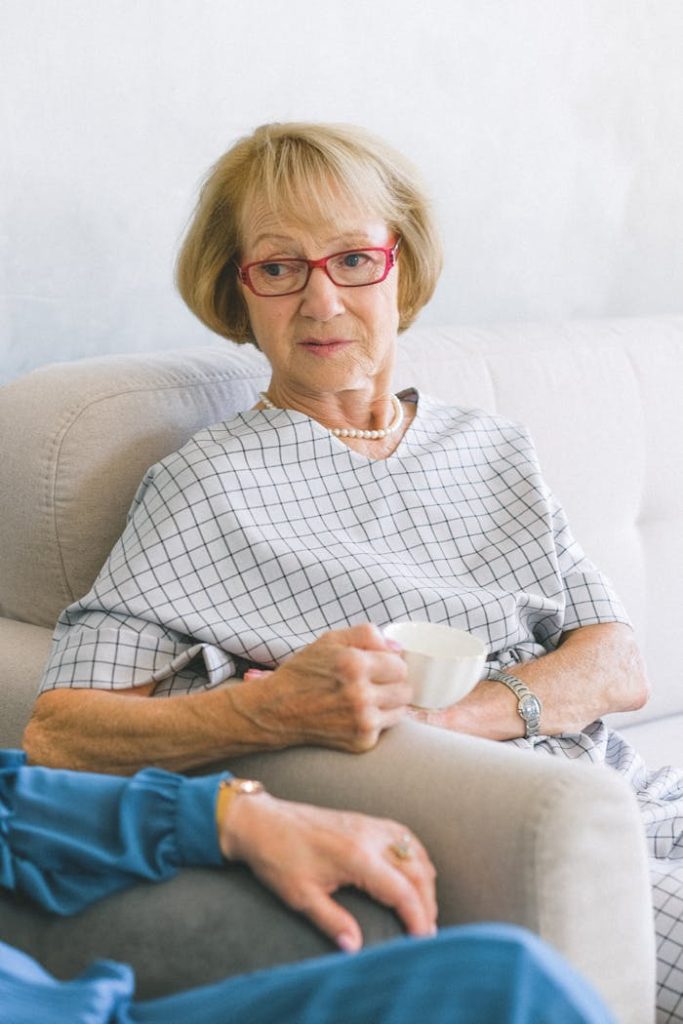 Senior female in eyeglasses and trendy dress drinking cup of coffee while speaking with crop anonymous friend