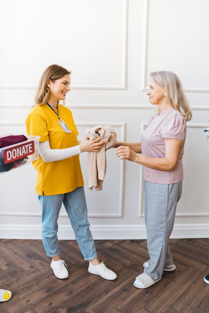 Smiling elderly woman receiving a sweater donation indoors from a young woman.