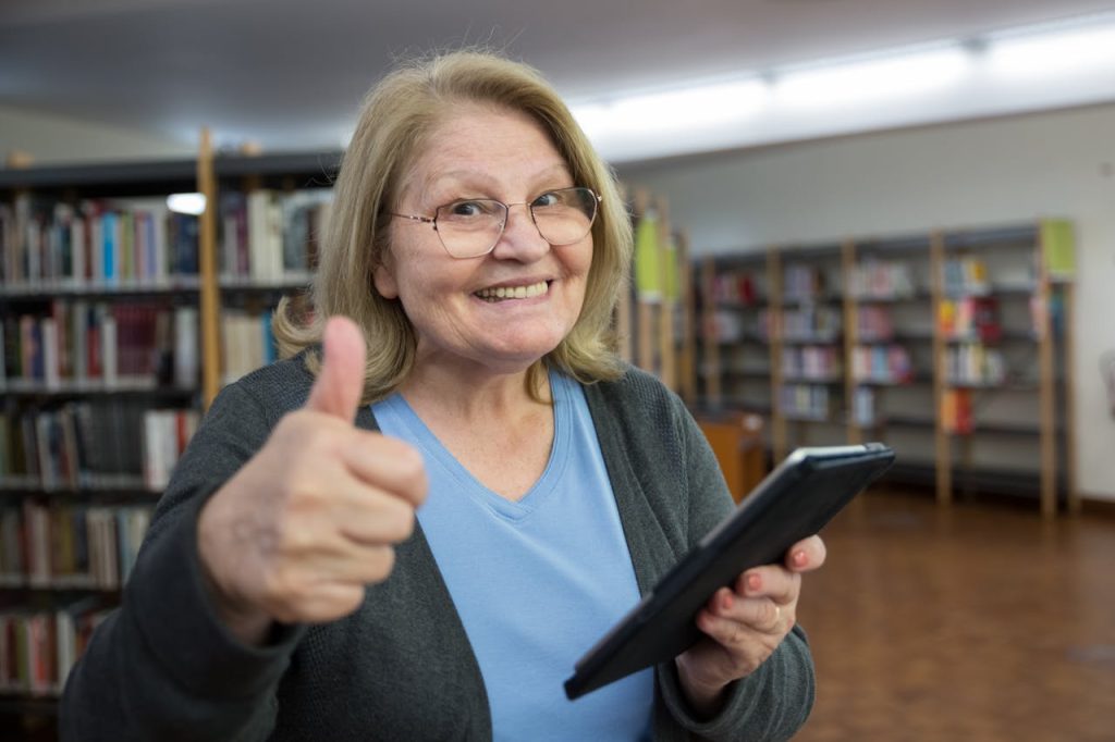 Happy senior woman gives a thumbs up while using a tablet in a library, embracing technology.