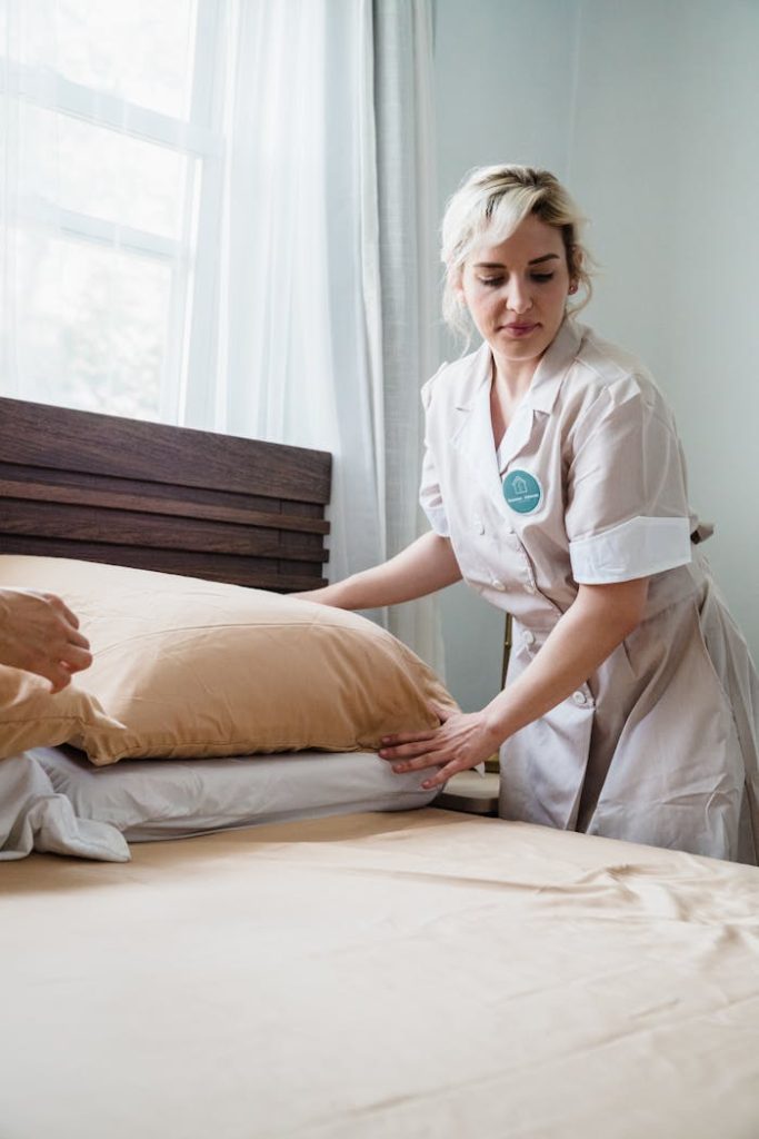 A female housekeeper tidily arranges pillows on a bed in a bright room.